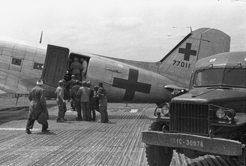 Wounded personnel being loaded onto a medical evacuation transport aircraft marked with a Red Cross, with a military ambulance parked in the foreground.