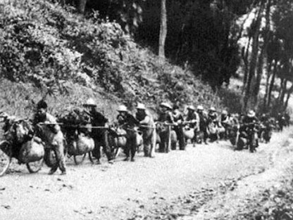 A line of Vietnamese supply workers pushing heavily loaded modified bicycles (xe đạp thồ) along a dirt road